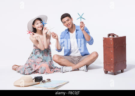 Couple in casual outfits holding starfish and sitting down on the floor with a suitcase and travel related materials Stock Photo