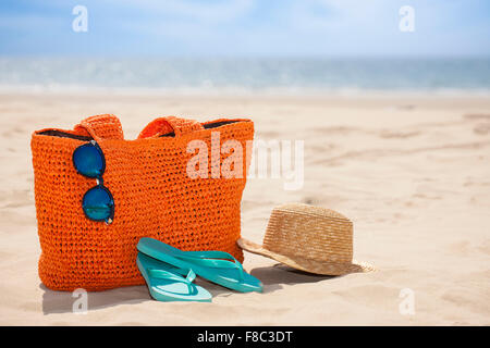 Sunglasses hanging on a bag, flip flops, and a hat on sand out of focus with the background of beach Stock Photo
