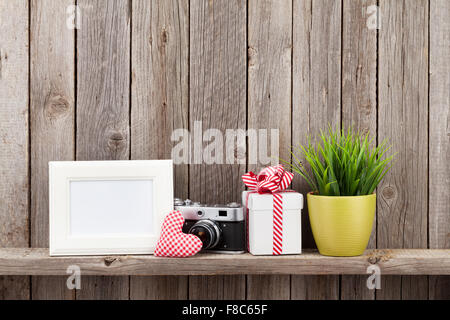 Blank photo frame, heart gift, camera and plant on shelf in front of wooden wall Stock Photo