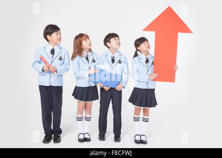 Four elementary school students standing with a big red pencil, a book, and a big sized arrow pointing up and all looking up Stock Photo