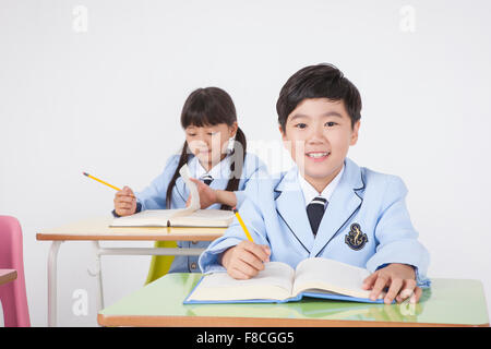 Elementary school boy in school uniforms seated at desk smiling with the background of a girl student seated at desk studying Stock Photo