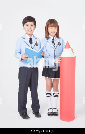 Elementary school boy standing with a book and a girl standing next to a big red pencil both in school uniforms and staring Stock Photo