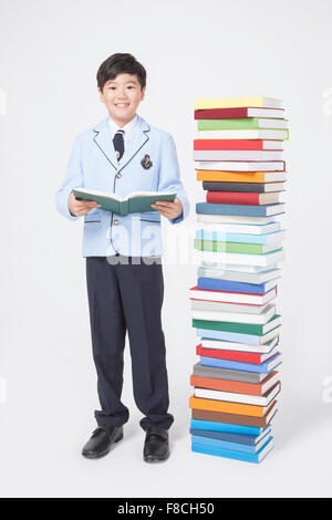 Elementary school boy in school uniforms holding a book and staring forward with a smile next to a pile of books Stock Photo