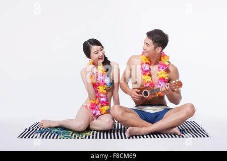 Couple in beach wear and flower necklaces sitting down on a towel and man playing ukulele both looking at each other and smiling Stock Photo
