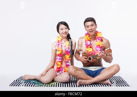Couple in beach wear and flower necklaces sitting down on a towel and man playing ukulele both staring forward and smiling Stock Photo