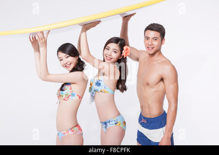 Two women in bikini and a man in swimming pants holding a surfing board above their heads all staring forward with a smile Stock Photo