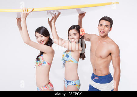 Two women in bikini and a man in swimming pants holding a surfing board above their heads all staring forward with a smile Stock Photo