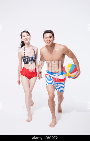 Man in swimming pants holding a beach ball running hand in hand with a woman in bikini both staring forward with a smile Stock Photo