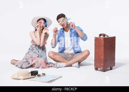 Man in casual outfits sitting on the floor with a woman in dress both holding starfish to cover their eyes with other travel Stock Photo