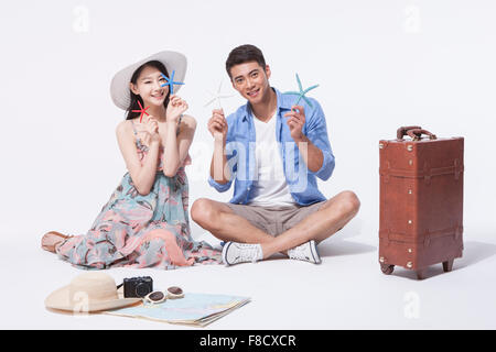 Man in casual outfits sitting on the floor with a woman in dress both holding starfish with other travel related objects around Stock Photo