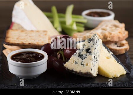 A cheese plate on slate with chutney, cheddar, stilton cheese, crackers, brie, grapes and celery. Stock Photo