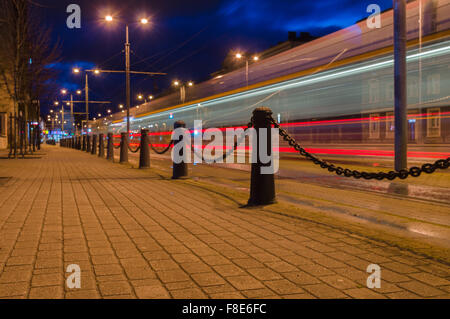 Trail of light left by tram at night. Paved street with decorative poles and chain Stock Photo