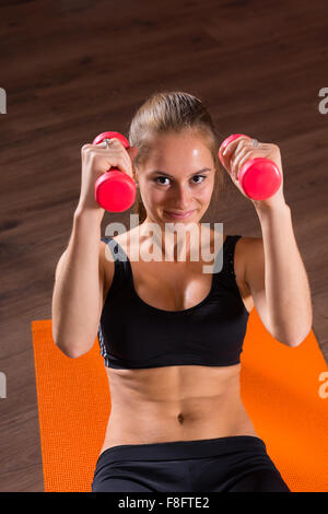 Portrait of Smiling Young Blond Woman Doing Ab Crunches with Hand Weights on Bright Orange Floor Mat and Looking at Camera in Exercise Studio. Stock Photo