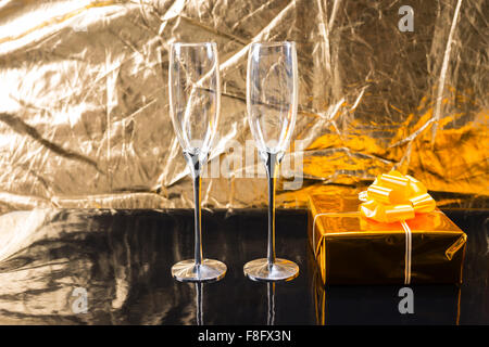 Pair of Empty Elegant Champagne Glasses on Shiny Black Table Beside Gift Wrapped in Gold Paper with Bow in front of Textured Metallic Gold Background. Stock Photo