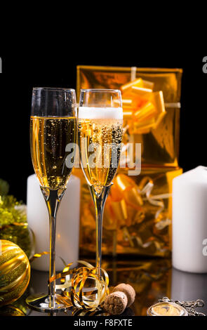 Festive Still Life - Pair of Glasses Filled with Sparkling Champagne in front of Black Background with Gold Wrapped Gifts, White Candles and Christmas Decorations. Stock Photo