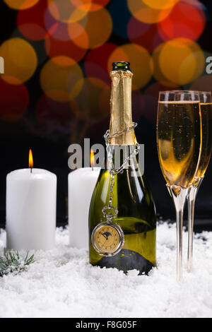 Two white burning candles near two stemmed crystal glasses and an open bottle of champagne with a vintage hanging clock on white snow arranged for the celebration of the New Year. Stock Photo