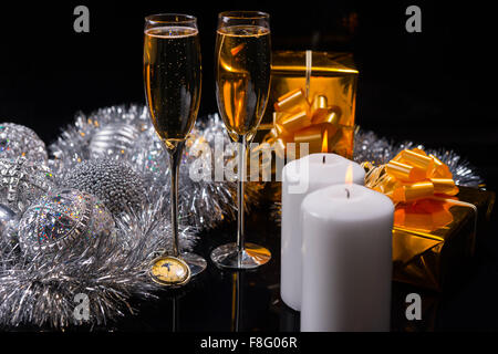 Festive Christmas Still Life - Pair of Glasses Filled with Sparkling Champagne on Table with Gifts, Silver Decorations and Lit White Pillar Candles on Black Surface. Stock Photo