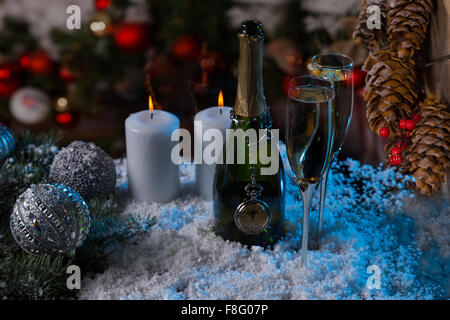 Festive Still Life - Bottle of Champagne with Two Full Glasses on Snowy Surface with Lit Candles and Surrounded by Christmas Decorations Illuminated by Blue Light. Stock Photo