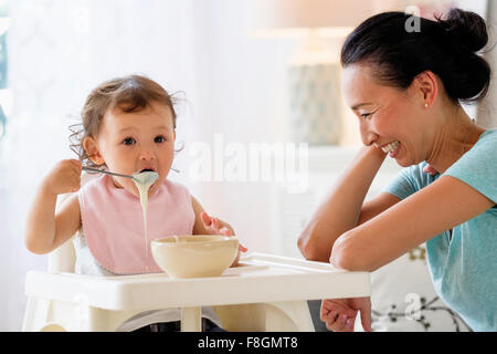 Mother watching baby daughter eat in high chair Stock Photo