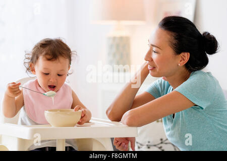 Mother watching baby daughter eat in high chair Stock Photo