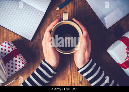 Christmas coffee, top view of female hands holding a cup of hot coffee drink on wooden table with Xmas gifts in boxes, retro ton Stock Photo