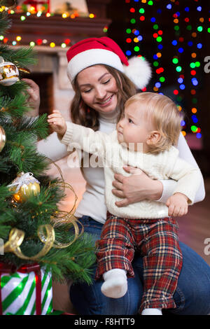 Mother in red santa hat looks at her little toddler son and smiling  near Christmas trees