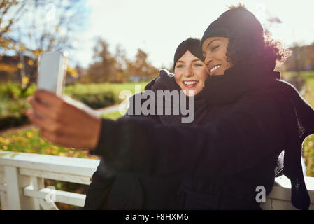 Two smiling young women in warm clothes taking selfie against of autumnal park Stock Photo