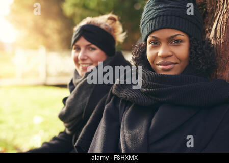 Close-up of two smiling young women leaning on tree trunk in autumnal park