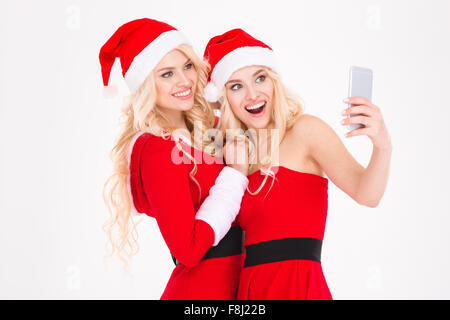 Cheerful blonde sisters twins in red santa claus dresses and hats making selfie using cell phone over white background Stock Photo