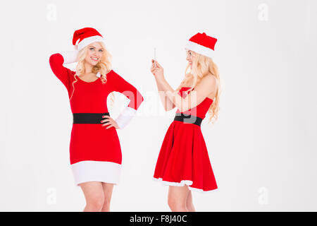 Two amusing beautiful sisters twins in red santa claus dresses and hats taking photos using cellphone over white background Stock Photo
