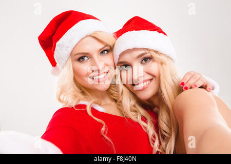 Selfie portrait of two lovely charming blonde sisters twins in santa claus costumes and hats isolated over white background Stock Photo