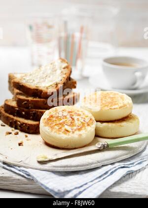 Crumpets and seeded wholemeal bread with butter on vintage chopping board with cup of tea Stock Photo