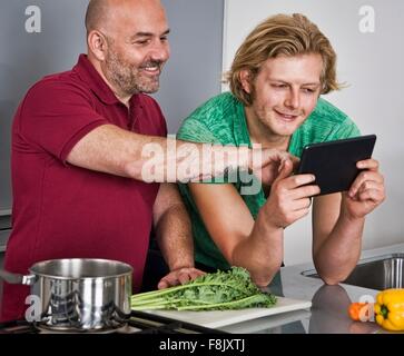 Male couple using digital tablet whilst preparing food in kitchen Stock Photo