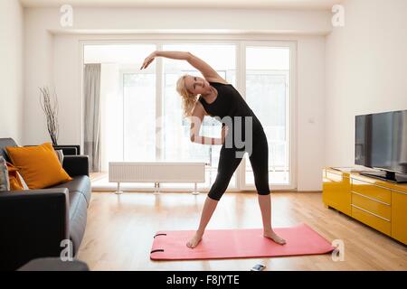 Mature woman in lounge standing on yoga mat bending over sideways arm raised stretching Stock Photo