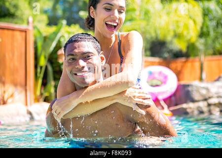 Mid adult man carrying young woman on back in swimming pool looking at camera smiling Stock Photo