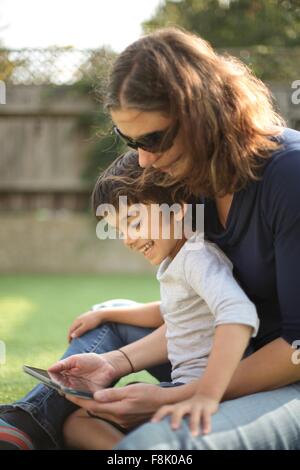 Side view of boy sitting on mother's lap using digital tablet looking down smiling Stock Photo