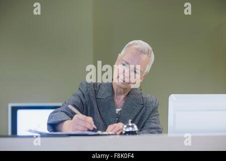 Senior female receptionist writing at hotel reception desk Stock Photo