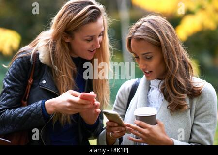 Two young female friends drinking coffee and reading smartphone texts in park Stock Photo