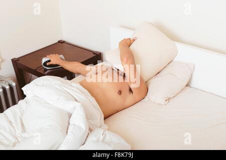 Young man in bed, covering face with pillow, turning off alarm clock Stock Photo