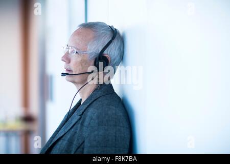 Side view of mature woman leaning against wall wearing telephone headset looking away Stock Photo