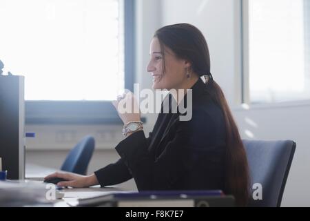 Side view of young woman in office sitting at desk using computer smiling Stock Photo