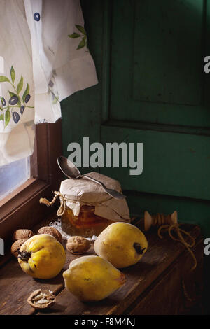 Three juicy quinces, walnuts and jar of honey over wooden table near window with bright sunlight. With green wooden wall at back Stock Photo