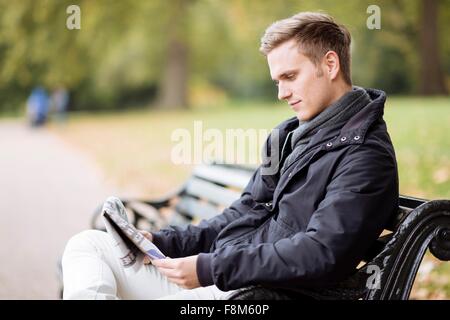 Young man sitting on bench reading newspaper in park Stock Photo