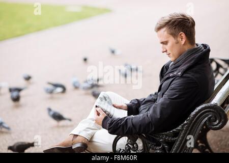 Young man sitting on park bench reading newspaper Stock Photo