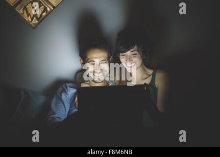 Front view of young couple illuminated by laptop computer screen, looking down smiling Stock Photo
