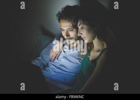 Young woman sitting with arm around young man illuminated by laptop computer screen Stock Photo