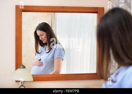 Pregnant woman holding stomach, standing in front of mirror Stock Photo