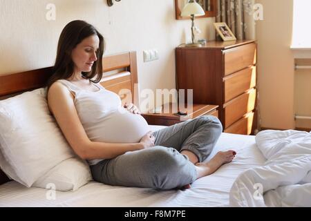 Pregnant woman sitting on bed, holding stomach Stock Photo