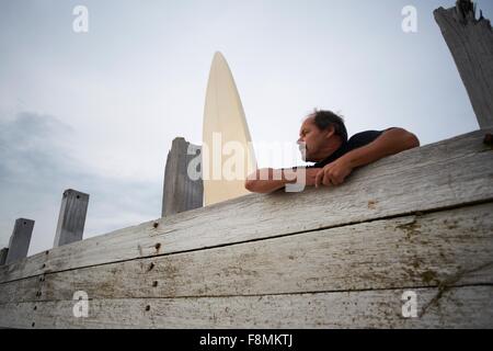 Surfer leaning against wooden fence with surfboard Stock Photo