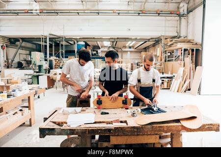 Front view of young men in carpentry workshop standing at workbench attaching wheels to skateboard Stock Photo